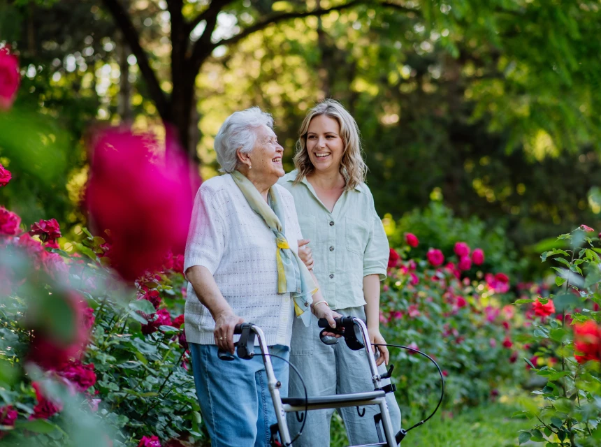 Junge Frau begleitet ältere Frau mit Rollator durch einen blühenden Rosengarten. Beide lachen und wirken gelöst.